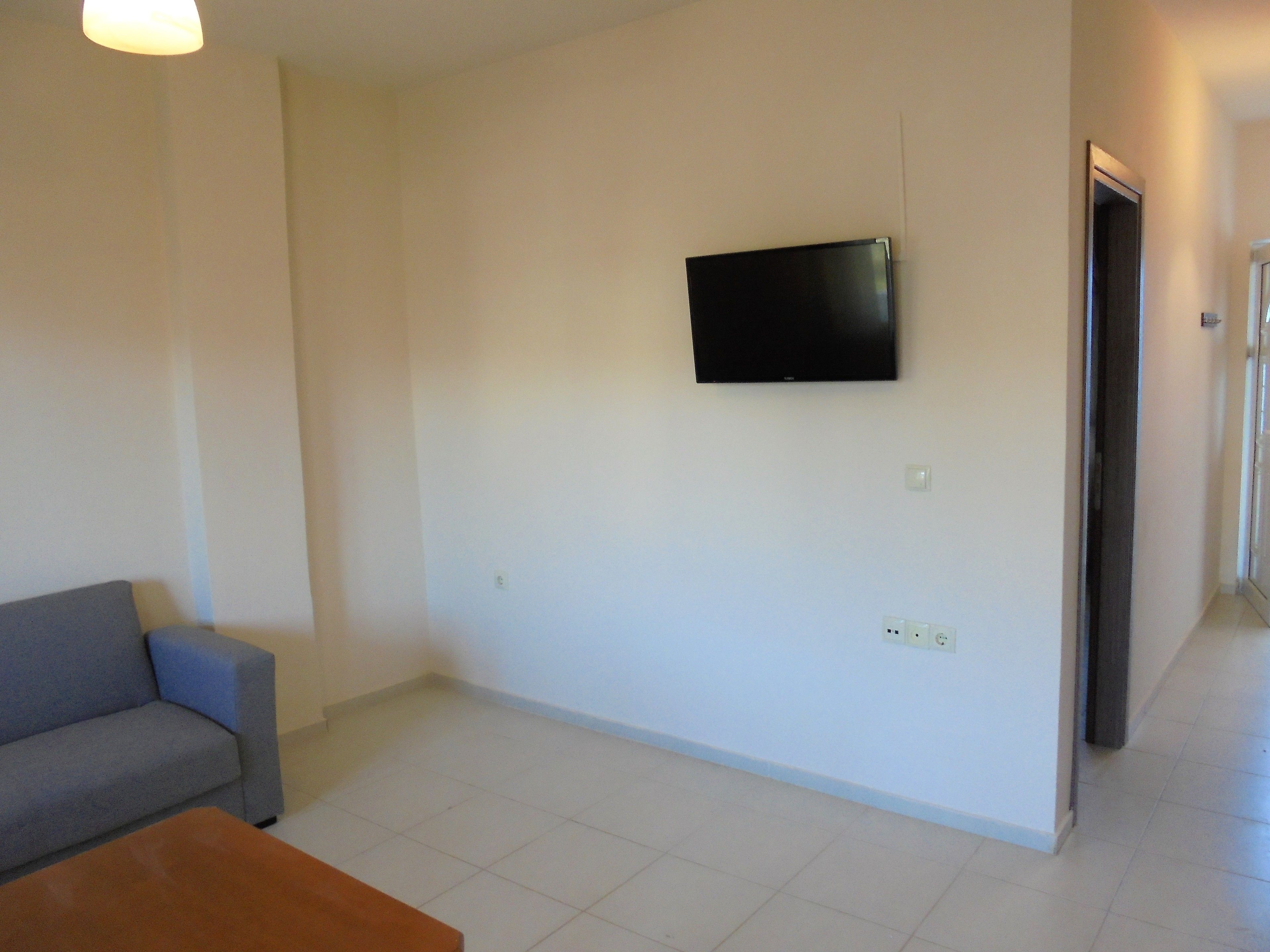 Una habitación con un sofá gris, una mesa de centro de madera, un televisor de pantalla plana montado en la pared y una puerta que da a otra habitación.