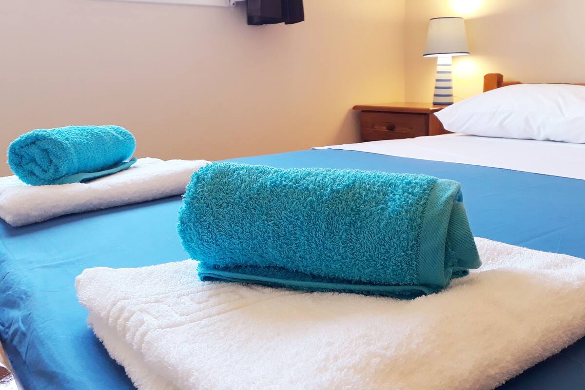 A hotel room with a neatly made bed, two rolled towels on the bed, a bedside lamp, and a nightstand.