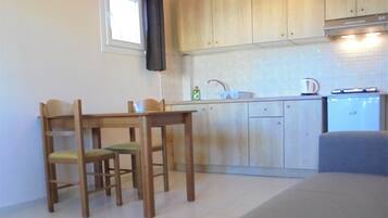 A compact kitchen with wooden cabinets, a dining table with chairs, a small refrigerator, and a window with a curtain.