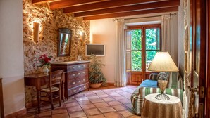 Una habitación acogedora con pared de piedra, vigas de madera en el techo, un aparador de madera, una silla, una mesa con una lámpara y una ventana con cortinas.