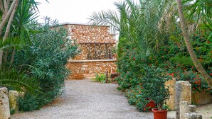 Un camino de grava que conduce a un edificio de piedra de aspecto rústico, rodeado de abundante vegetación y plantas en macetas.