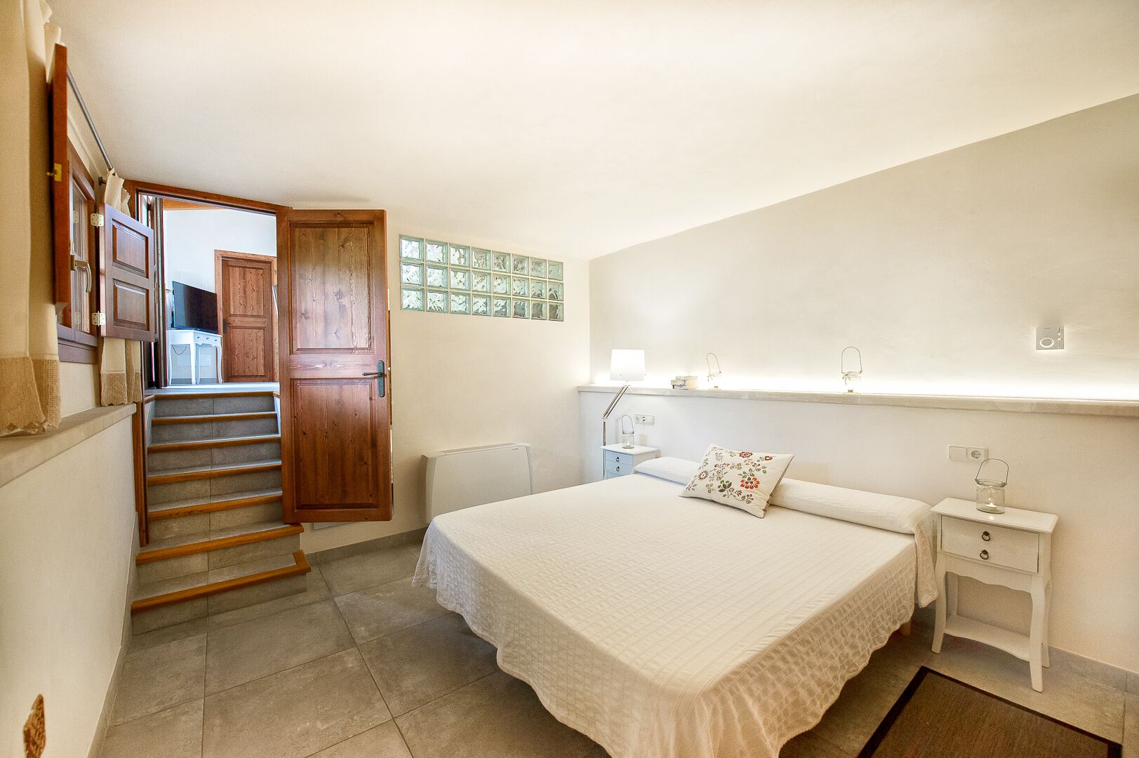 A bedroom with a bed, bedside table, nightstand, lamp, and a wooden door leading to a kitchen.