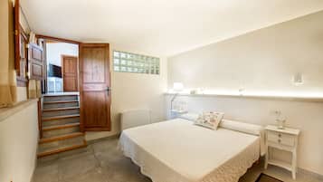 A bedroom with a bed, bedside table, nightstand, lamp, and a wooden door leading to a kitchen.
