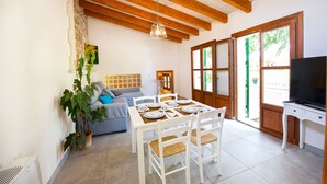 Un comedor con una mesa dispuesta para cuatro personas, un sofá y un televisor en una habitación con vigas de madera y ventanas amplias.