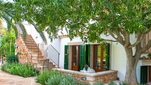 Una casa con una escalera de piedra, contraventanas verdes y un árbol que da sombra.