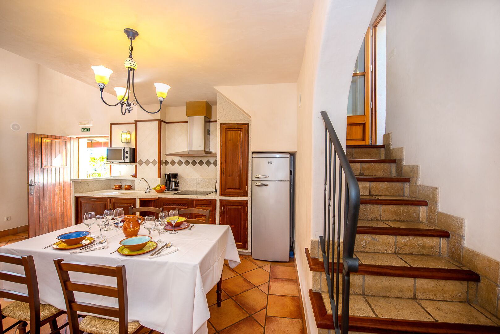 Un coin repas avec une table dressée pour quatre personnes, une cuisine équipée d’un réfrigérateur et un escalier.
