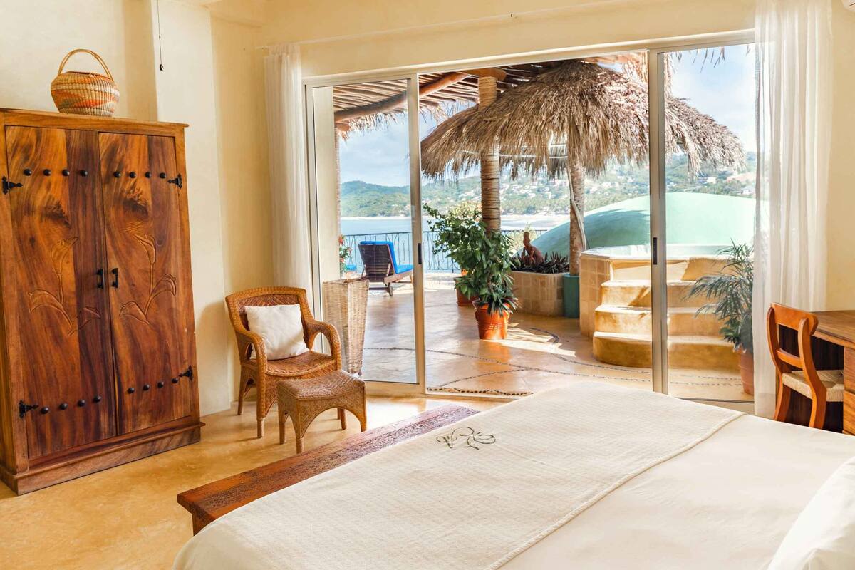 A bedroom with a large bed, a wooden wardrobe, a wicker chair, and a view of a balcony with a thatched roof and plants.