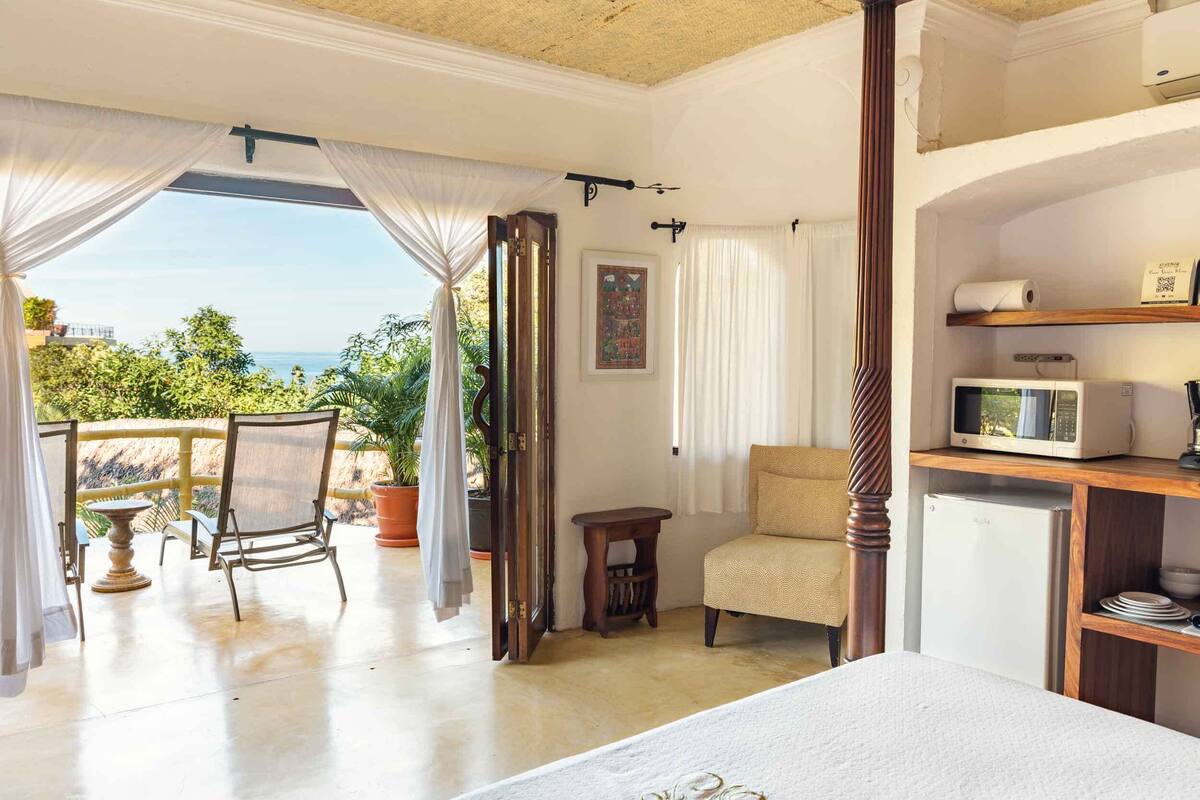 A bedroom with a bed, a chair, a small table, a microwave, and a view of the sea.