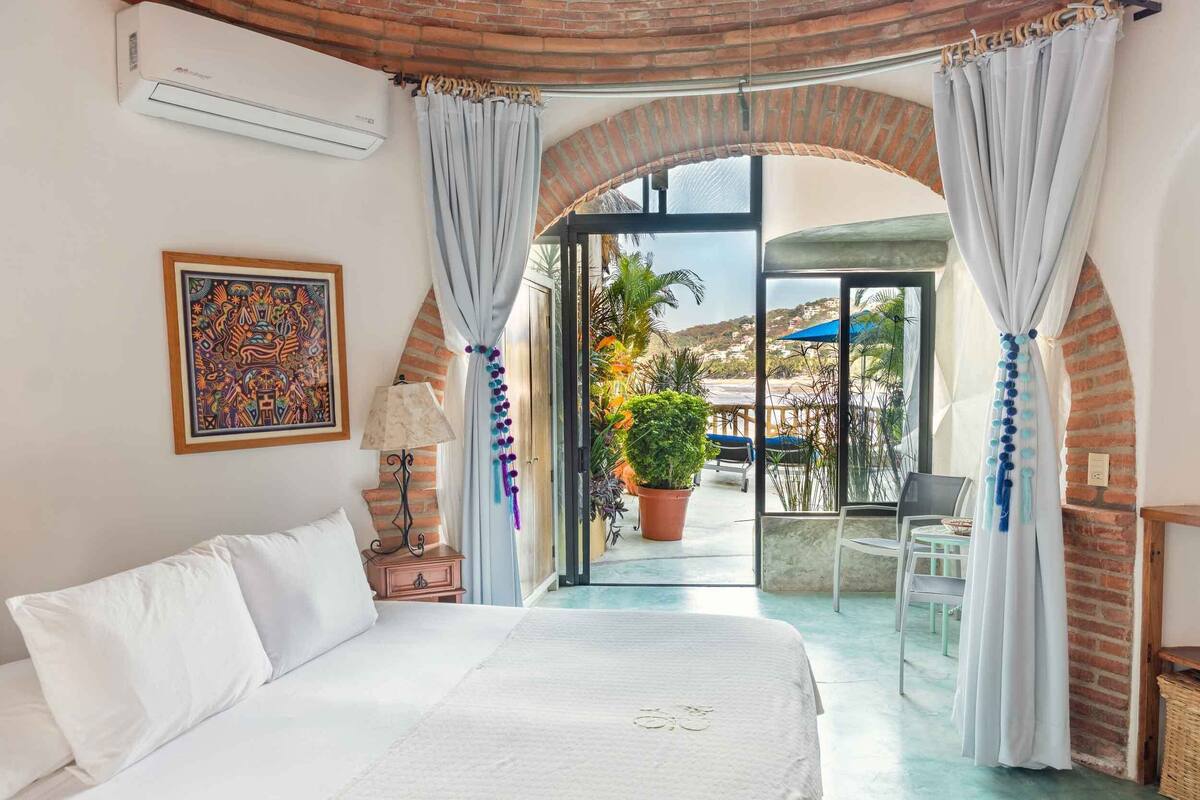 A bedroom with a large bed, a nightstand, a lamp, and a view of the balcony with plants and a chair.