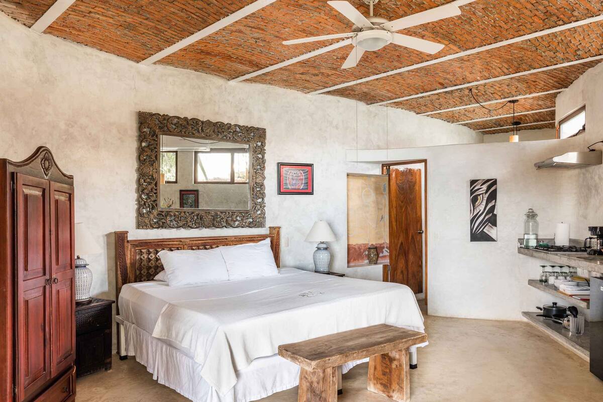A bedroom with a large bed, a wooden bench, a ceiling fan, a mirror, and a kitchenette.