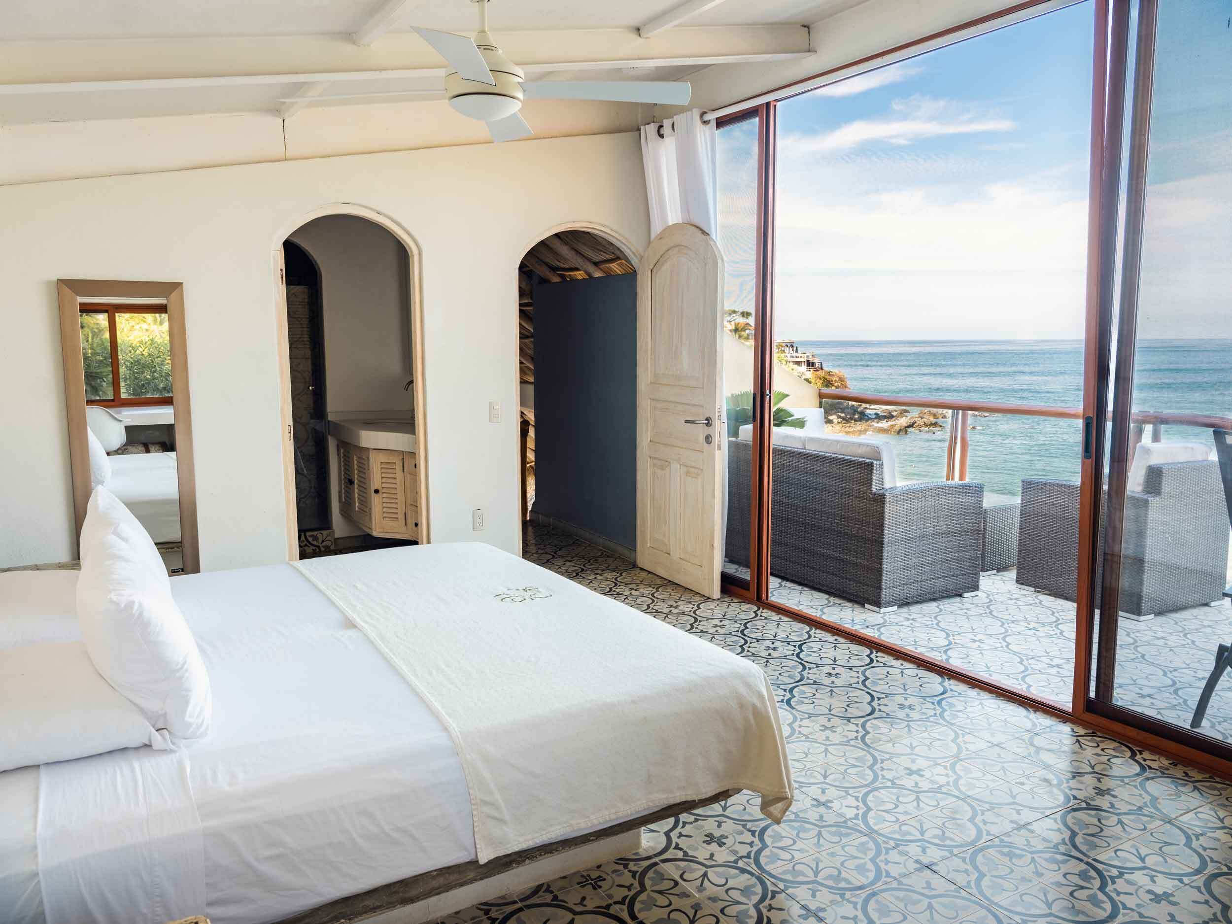A hotel room with a bed, a ceiling fan, a large window with a view of the sea, and a balcony with wicker furniture.