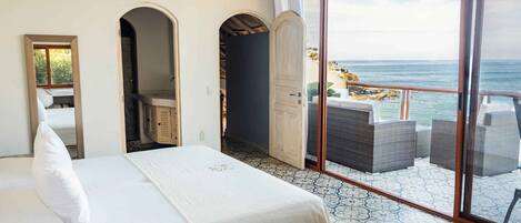 A hotel room with a bed, a ceiling fan, a large window with a view of the sea, and a balcony with wicker furniture.