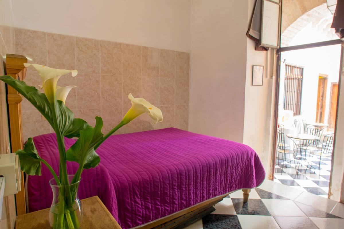 Une pièce comprenant un lit, un vase avec des fleurs et donnant sur un espace extérieur aménagé avec des chaises et des tables.