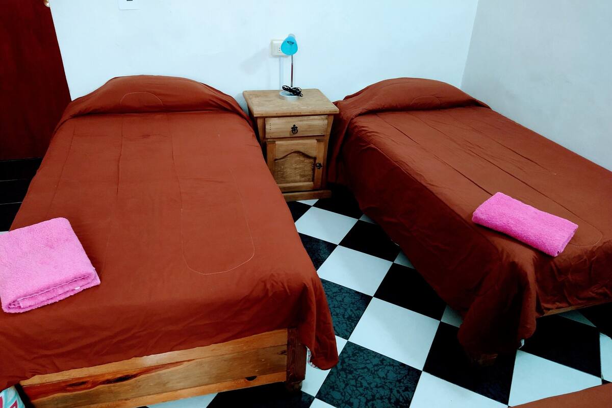 Une chambre avec deux lits, chacun agrémenté d’une serviette rose, une table de chevet en bois avec une lampe, et un sol à carreaux.