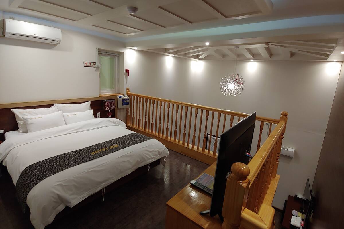 A hotel room with a bed, a desk with a computer, a TV, a wooden railing, and an air conditioning unit.