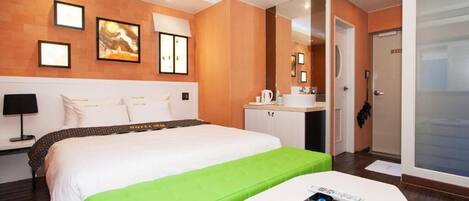 Quarto de hotel com uma cama grande, um banquinho verde estofado, uma mesa pequena branca, uma escrivaninha com um computador e um banheiro com pia e espelho.
