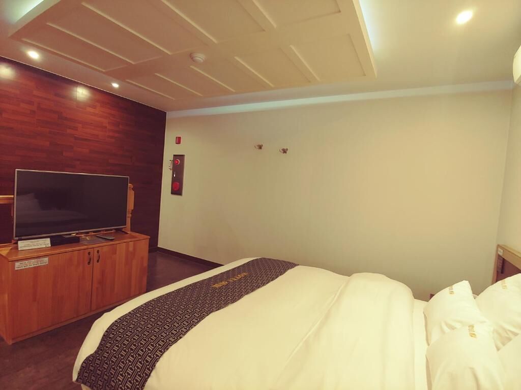 A hotel room with a bed, a TV on a wooden cabinet, and a plain wall with a fire alarm.