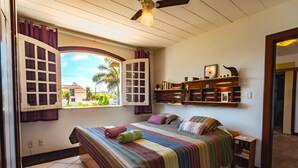 Ein Schlafzimmer mit großem Fenster, einem Bett mit gestreifter Bettwäsche, einem Deckenventilator und einem hölzernen Regal mit Büchern und Dekoration.