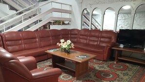 A living room with a brown leather sofa, a wooden coffee table with a vase of flowers, and a television on a wooden stand. - Kok-Tobe Almaty Hotel (Almaty)