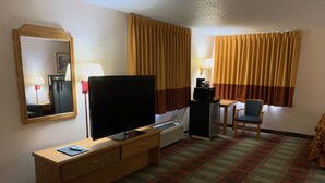 A hotel room with a flat-screen TV, a desk with a lamp, a chair, a bed with a patterned cover, and a large mirror.
