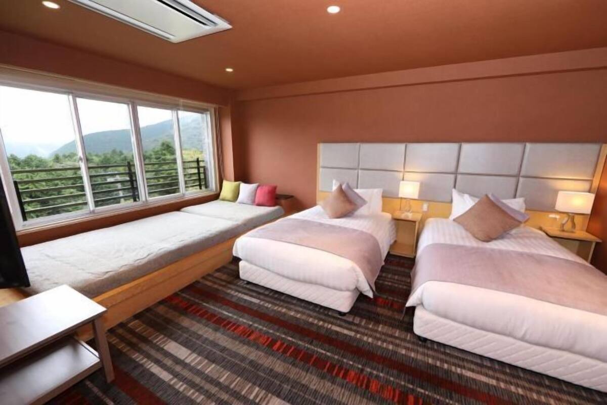 A hotel room with two beds, a bench, a small table, and a large window with a mountain view.