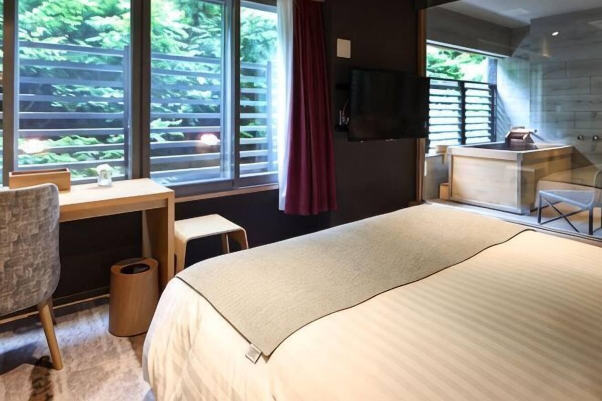 A hotel room with a bed, a desk, a chair, a TV, and a balcony with a view of greenery.