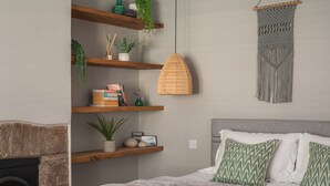 A bedroom with a bed, a fireplace, wooden shelves with plants and books, a woven pendant light, and a macramé wall hanging. - Seascape Hideaways at Mortehoe (Woolacombe)