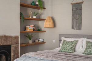 Un dormitorio con cama, chimenea, estanterías de madera con plantas y libros, una lámpara colgante de fibra natural y un tapiz de macramé.
