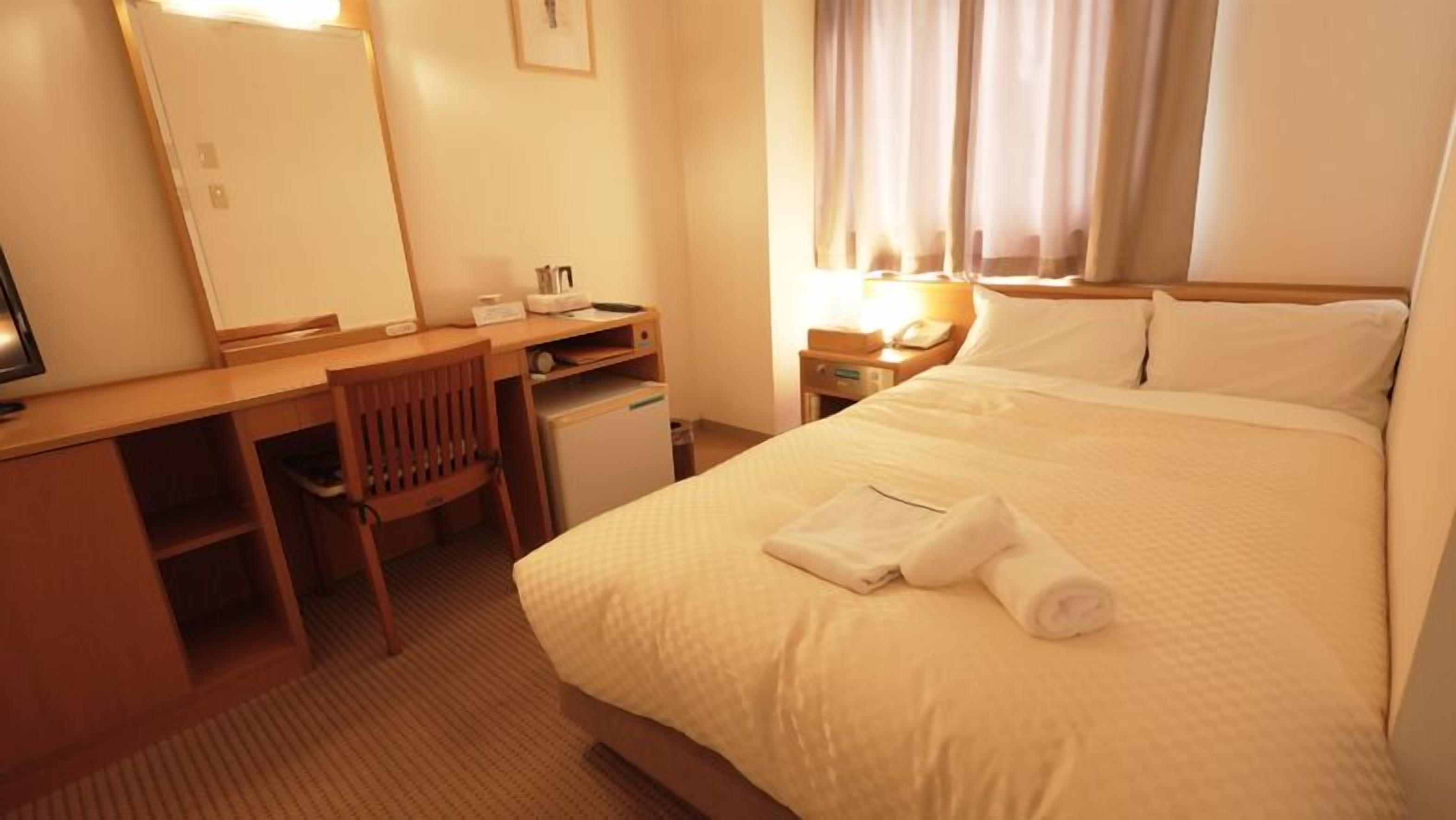 Ein Hotelzimmer mit einem Bett, einem Schreibtisch, einem Stuhl, einem Spiegel, einem Nachttisch und einem Fenster mit Vorhängen.