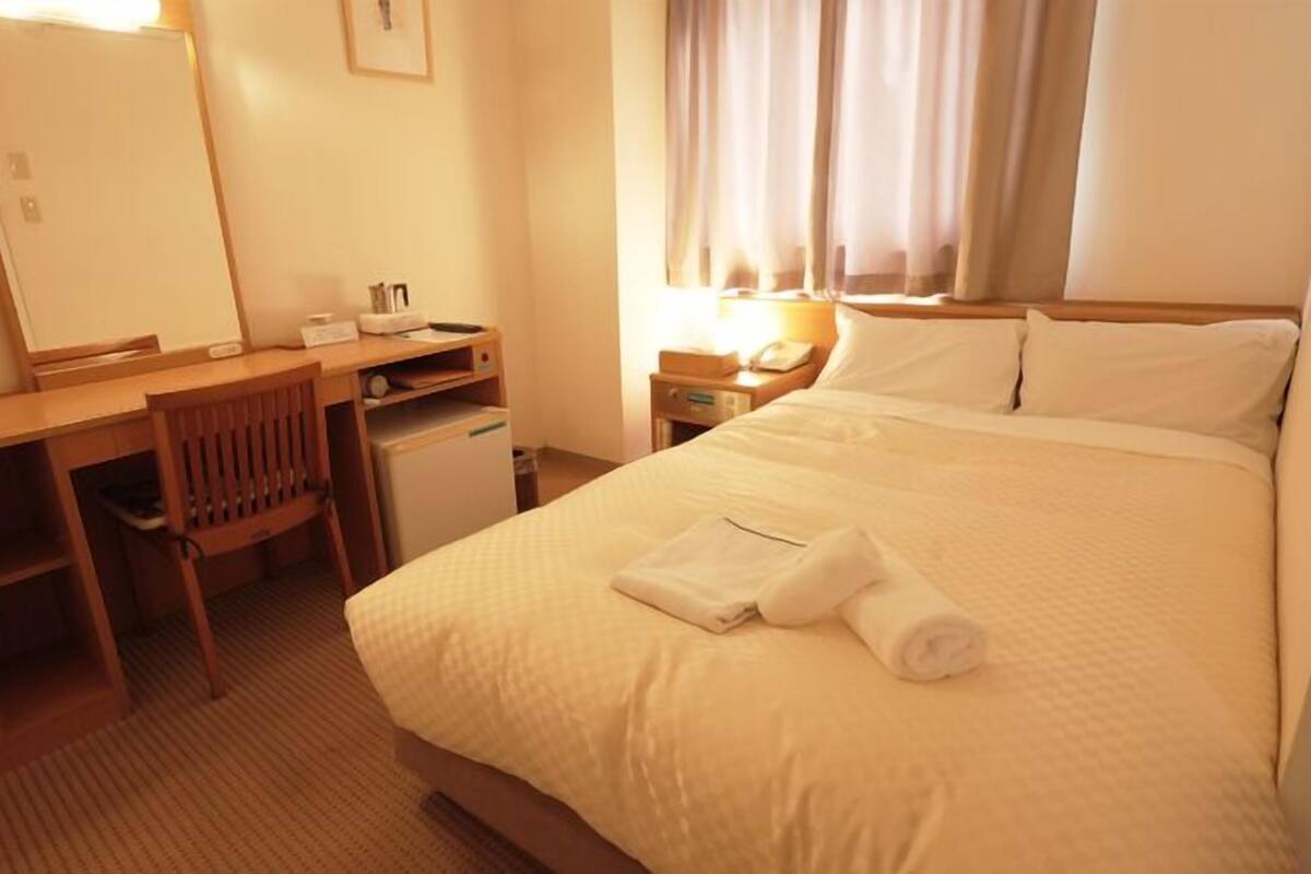 Ein Hotelzimmer mit einem Bett, einem Schreibtisch, einem Stuhl, einem Spiegel, einem Nachttisch und einem Fenster mit Vorhängen.