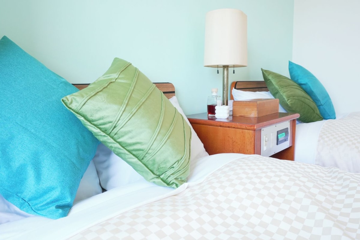 A hotel room with a bed, pillows, a bedside table with a lamp, and a digital clock.