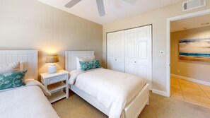A bedroom with two beds, a nightstand, a lamp, a door, and a painting on the wall. - House Of The Sun #315GF (Sarasota)