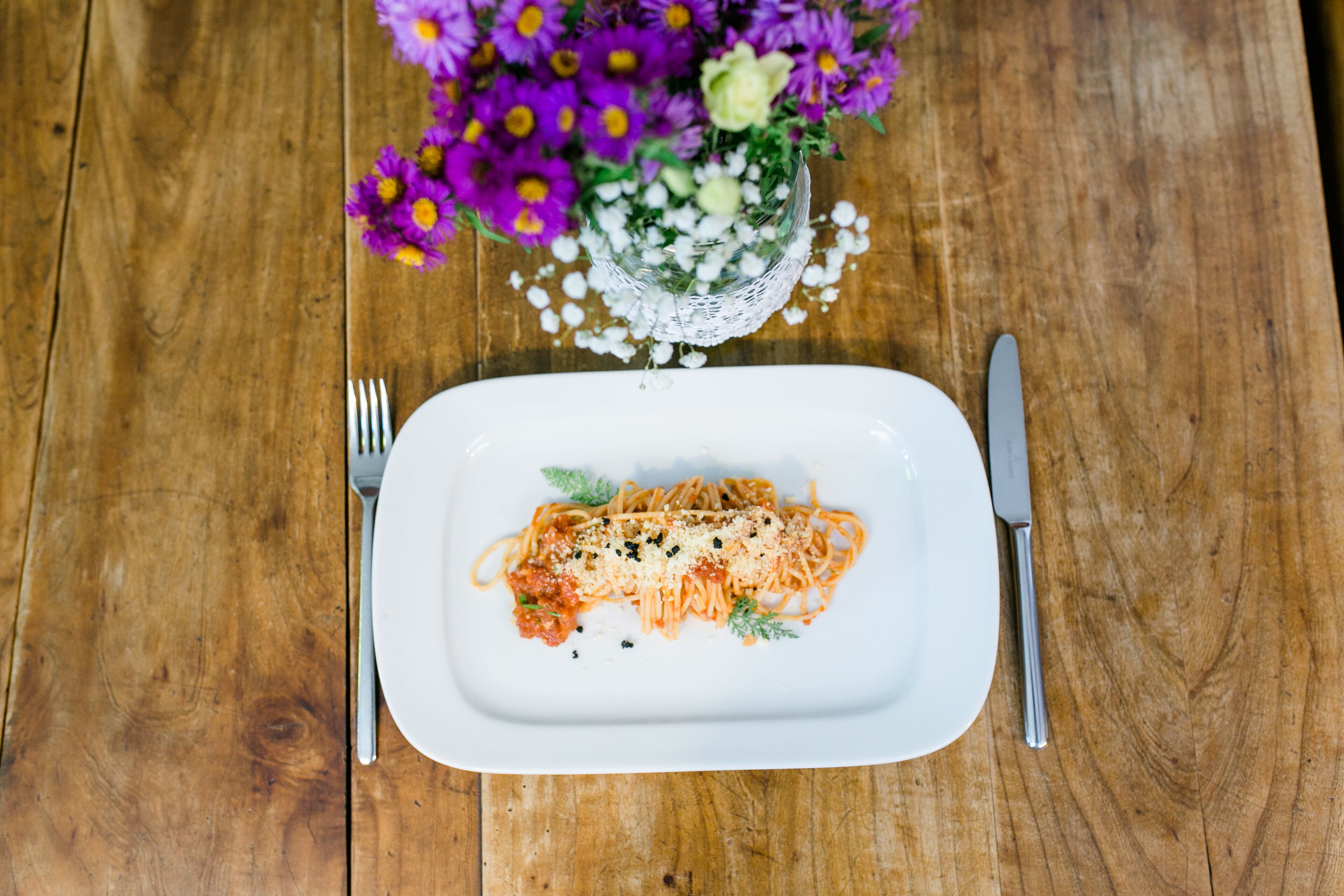 Une assiette de spaghettis à la sauce tomate et au fromage, une fourchette, un couteau et un vase de fleurs sur une table en bois.