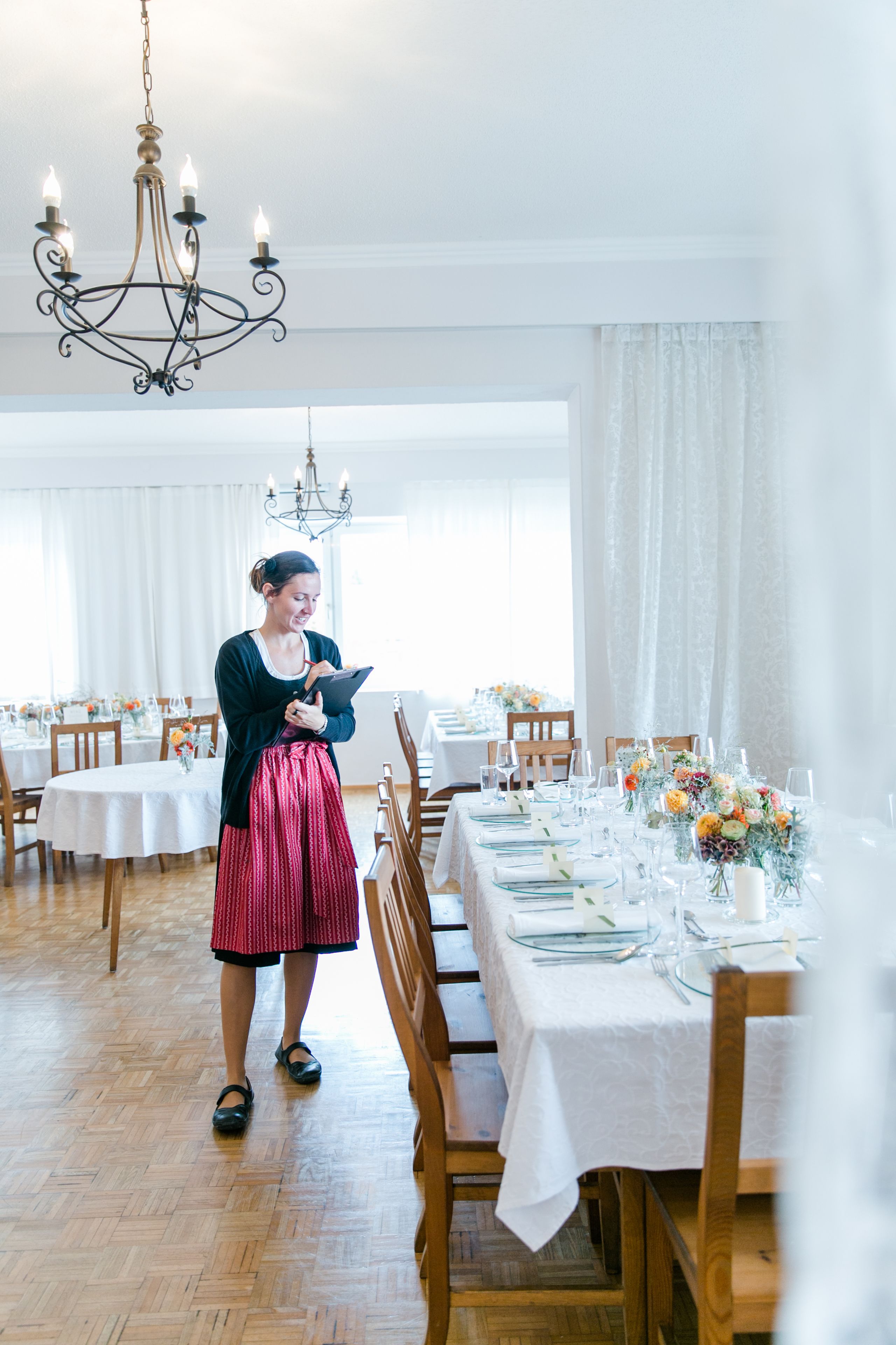 A dining area with tables set for a meal, a chandelier, and a person standing by the tables.