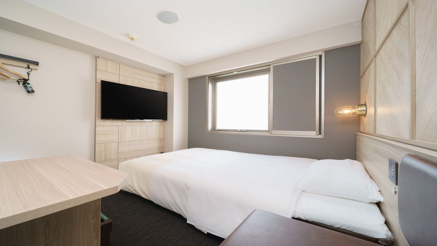 A single bed with a headboard, a flat-screen TV mounted on a wooden panel wall, a bedside table, and a window with a view.