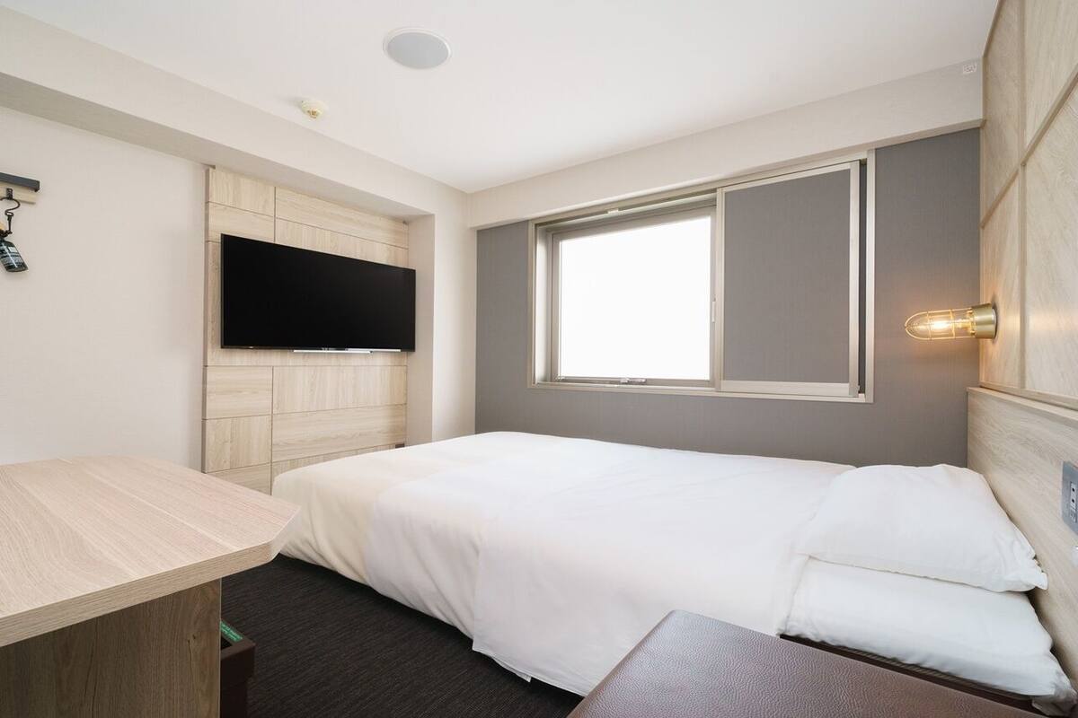 A single bed with a headboard, a flat-screen TV mounted on a wooden panel wall, a bedside table, and a window with a view.