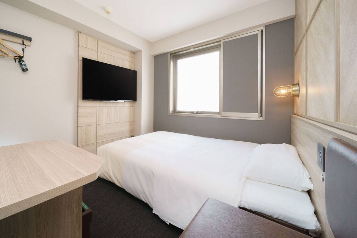A compact hotel room with a bed, a small desk, a mounted TV, and a window with a view.