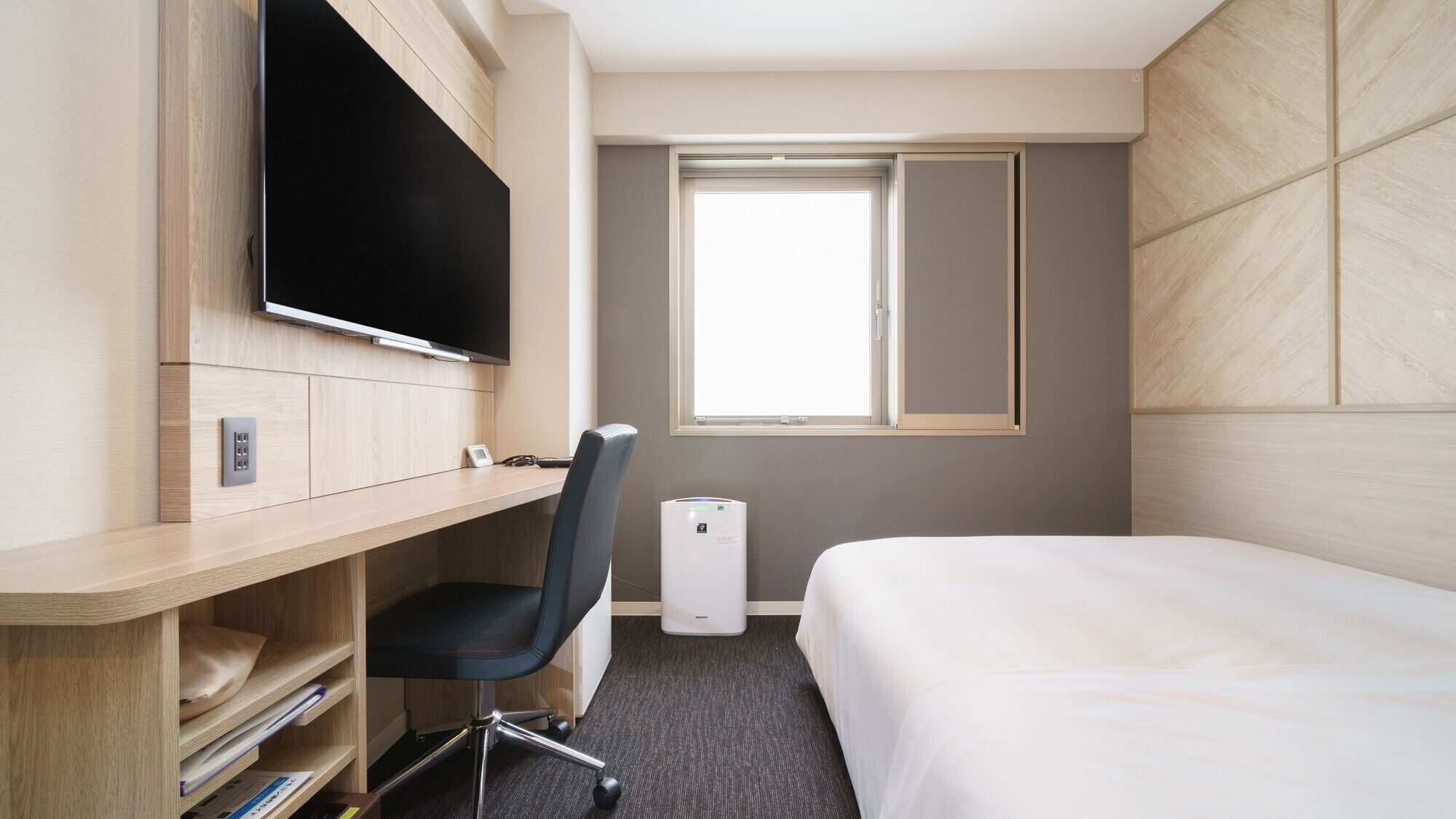 A hotel room with a bed, a desk with a TV, a chair, a window, and a thermostat.