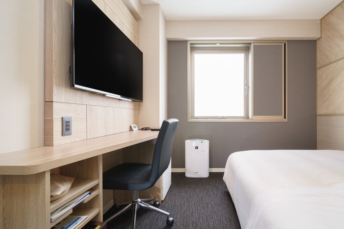 A hotel room with a bed, a desk with a TV, a chair, a window, and a thermostat.