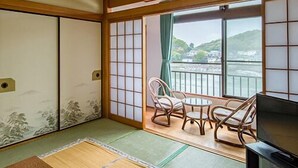 Una habitación japonesa tradicional con suelo de tatami, puertas correderas, televisor y balcón con vistas a una colina.