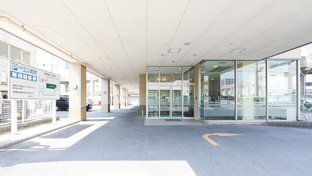 Entrada de un edificio con un camino señalizado en el suelo, puerta de cristal y un cartel a la izquierda.