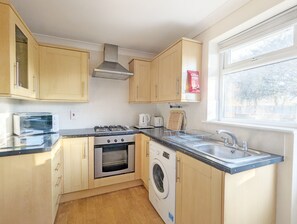 Een compacte keuken met houten kasten, een inbouwoven, een wasmachine en een raam met uitzicht op bomen.