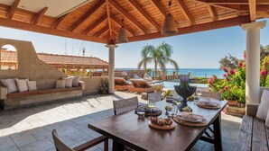 An outdoor patio with a wooden table set for dining, a cushioned seating area, and a view of the sea. - Villa Del Nonno (Rhodes)