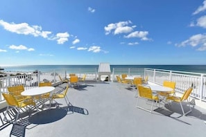 An outdoor seating area with yellow chairs and tables, a white railing, and a view of the ocean.