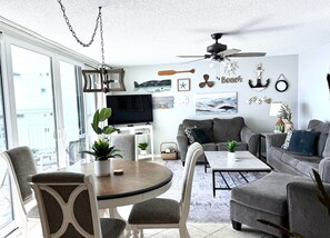 A modern living room with a round dining table, grey sofas, a ceiling fan, and nautical decor.
