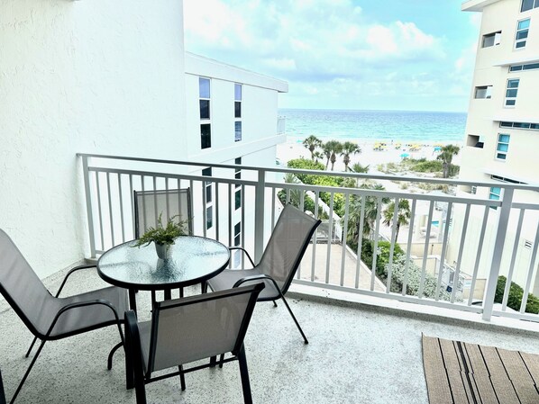 A balcony with a round glass table and two chairs, overlooking a beach with palm trees and a clear sky.