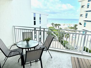 Varanda com mesa de vidro redonda e duas cadeiras, com vista para uma praia com palmeiras e céu claro.