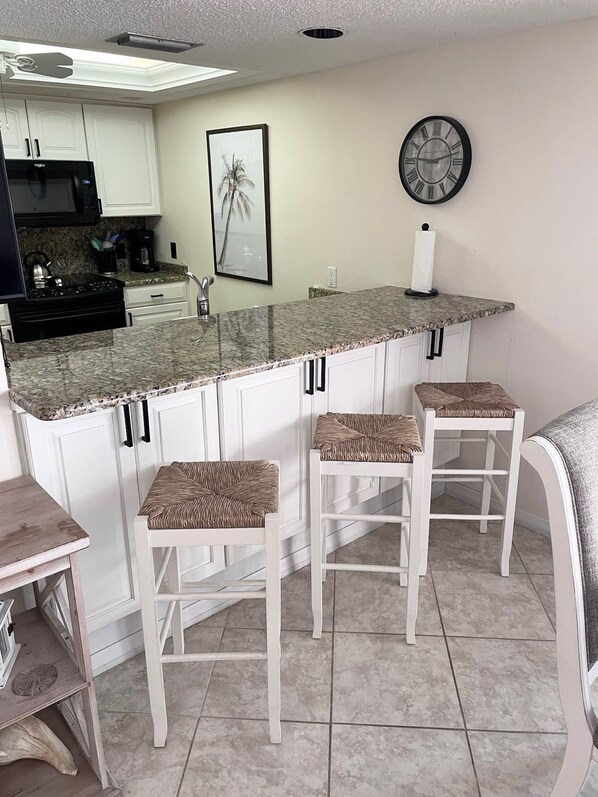 A kitchen with a marble countertop, white cabinets, a clock, and three wicker bar stools.