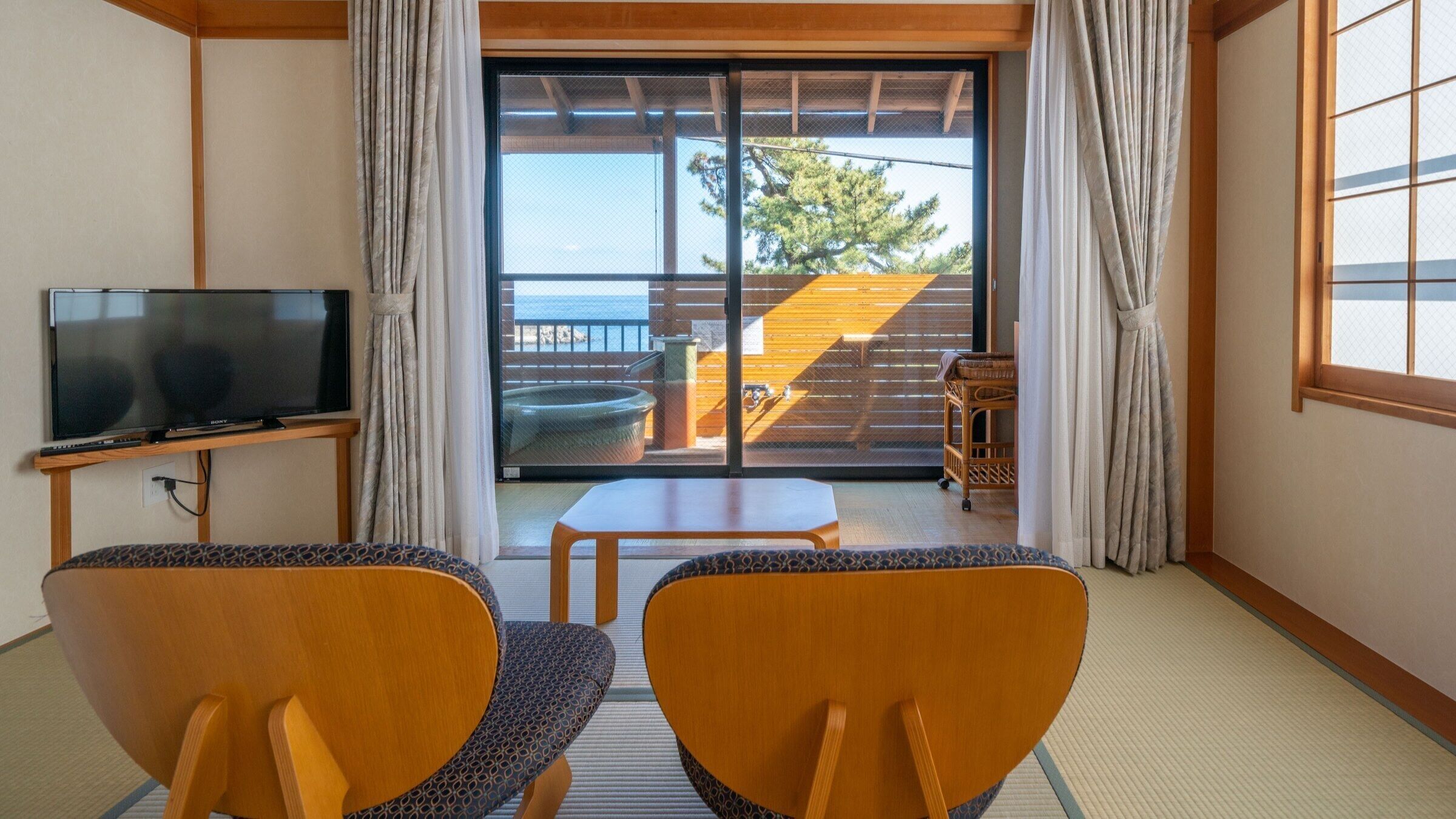 A room with a TV, two chairs, a small table, and a sliding glass door leading to an outdoor area with a wooden deck and a view of trees.