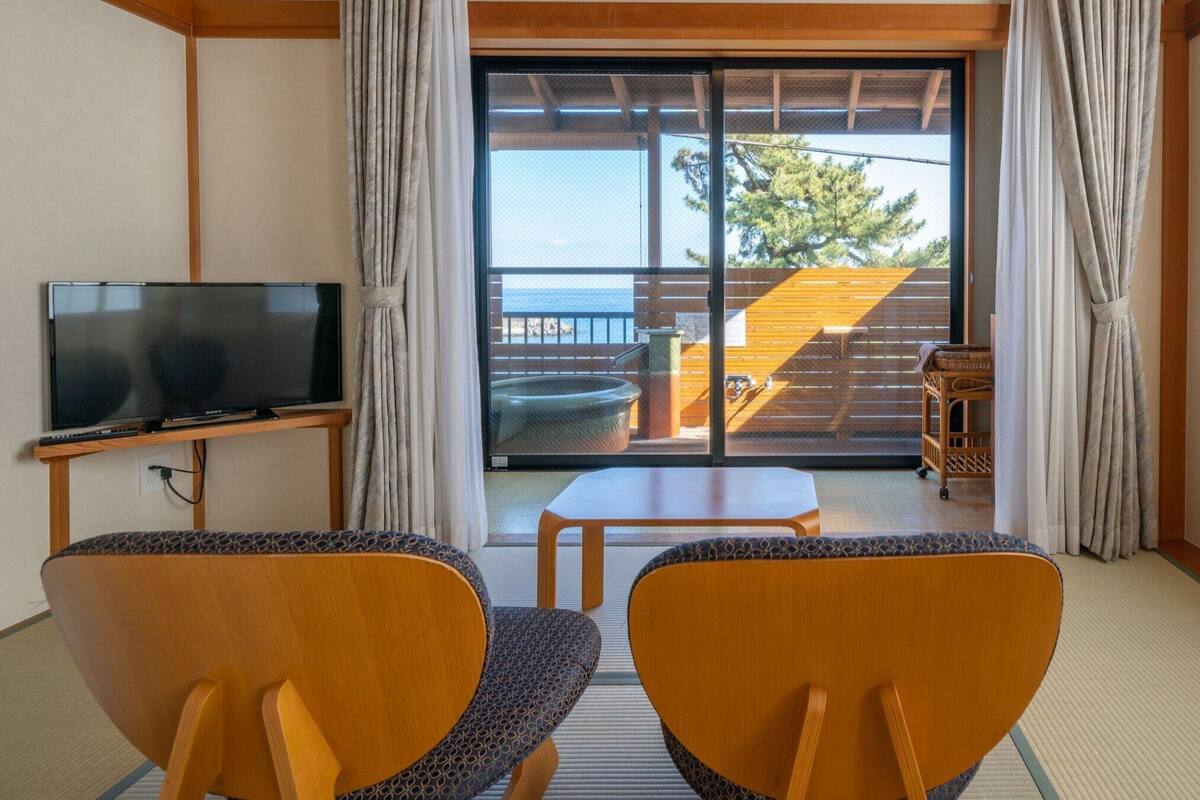 Quarto com televisão, duas cadeiras, uma mesinha e porta de vidro deslizante para área externa com deck de madeira e vista para árvores.